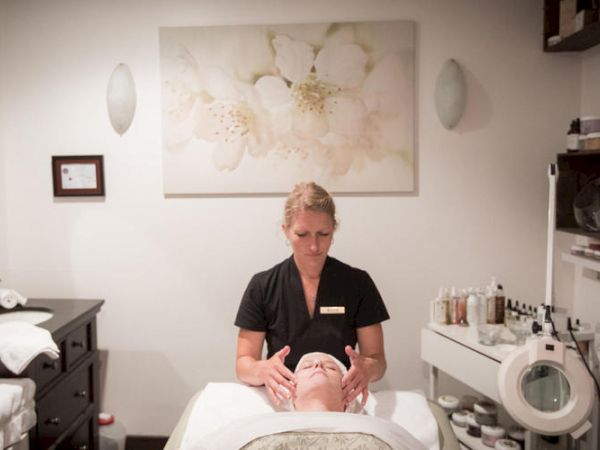 A woman receives a facial treatment from a therapist in a serene spa room decorated with floral art and skincare products on shelves.