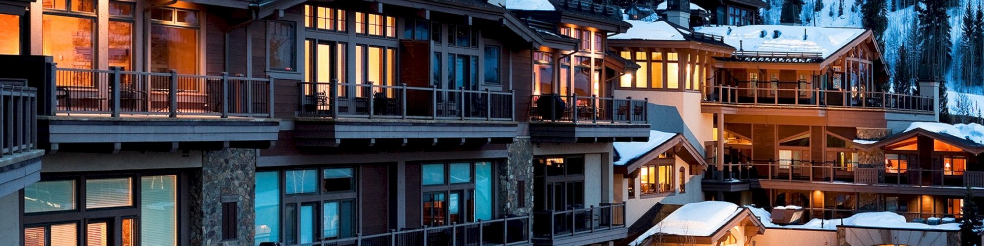 The image shows a series of lit-up buildings with balconies, situated against a snowy mountainous backdrop, giving a cozy, winter resort vibe.