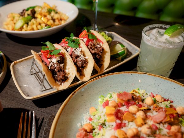 The image shows a table with beef tacos, a salad, a pasta dish, and a margarita garnished with a lime wedge, all on a dark surface.