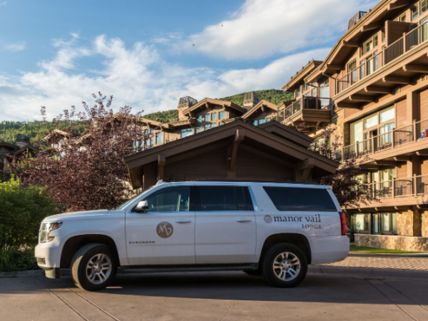 The image shows a white vehicle with "Manor Vail Lodge" parked in front of a large building complex with balconies, set against a mountainous backdrop.