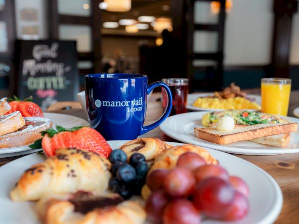 A table set with a variety of breakfast foods, including pastries, fresh fruits, toast, juice, and a mug labeled "manor vial hotel."