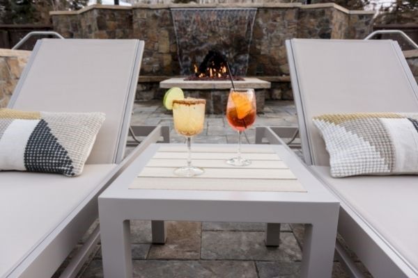 Two lounge chairs with pillows flank a small table holding two cocktails, set in front of an outdoor fireplace.