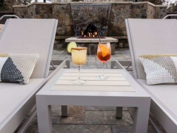 Two lounge chairs with pillows flank a small table holding two cocktails, set in front of an outdoor fireplace.