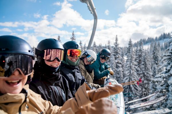 A group of people in ski gear riding a ski lift, smiling against a snowy forest backdrop and a clear blue sky.