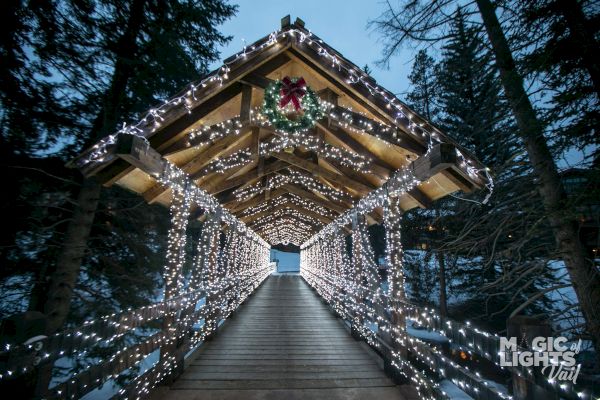 A covered wooden bridge adorned with white string lights and a wreath, set in a snowy forest. Branded 