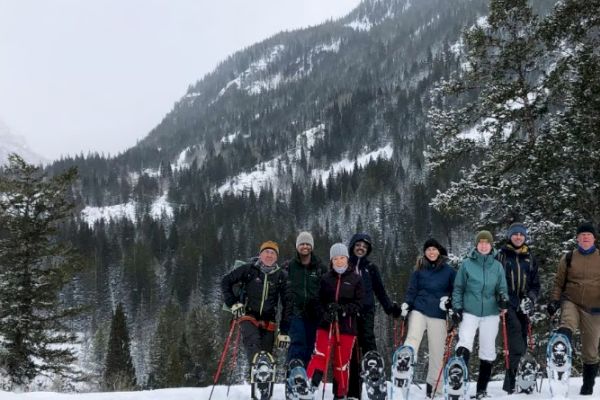 A group of people with snowshoes stands in a snowy mountain landscape, surrounded by trees.