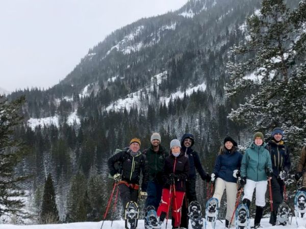A group of people with snowshoes stands in a snowy mountain landscape, surrounded by trees.