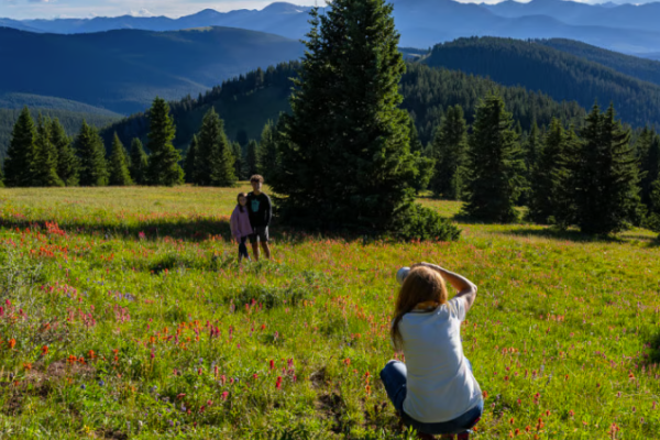 A person is photographing two people in a colorful mountain meadow with tall evergreen trees and distant hills in the background.