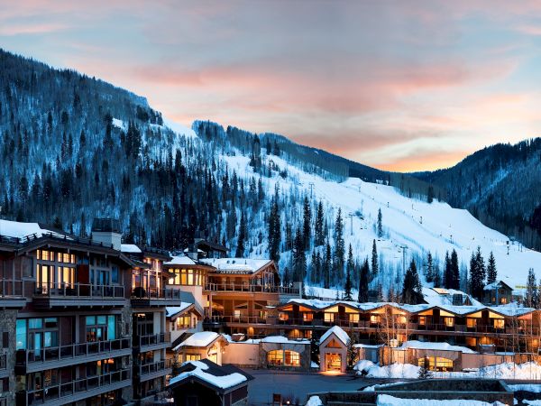 A snowy mountain resort with buildings in the foreground, ski slopes in the background, and a colorful sky at sunset.