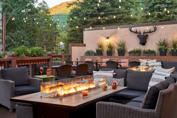 An outdoor seating area with cozy couches, a fire table, string lights, and potted plants, set against a backdrop of trees and mountains.