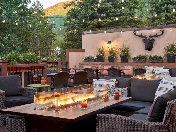An outdoor seating area with cozy couches, a fire table, string lights, and potted plants, set against a backdrop of trees and mountains.