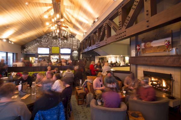 A bustling restaurant with people dining, cozy seating by a fireplace, and a bar with chalkboard art on the wall. Warm lighting fills the space.