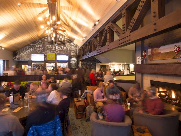 A bustling restaurant with people dining, cozy seating by a fireplace, and a bar with chalkboard art on the wall. Warm lighting fills the space.