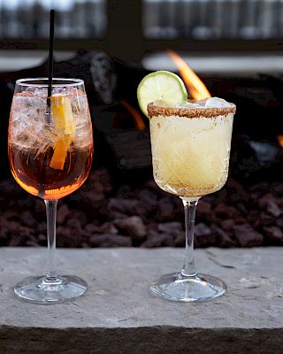 Two cocktails are on a stone surface: one with an orange slice and straw, the other with a lime wedge and salted rim, with a fire pit behind.