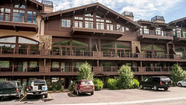 The image shows a multi-story rustic building with wooden balconies and parked cars in front under a partly cloudy sky.