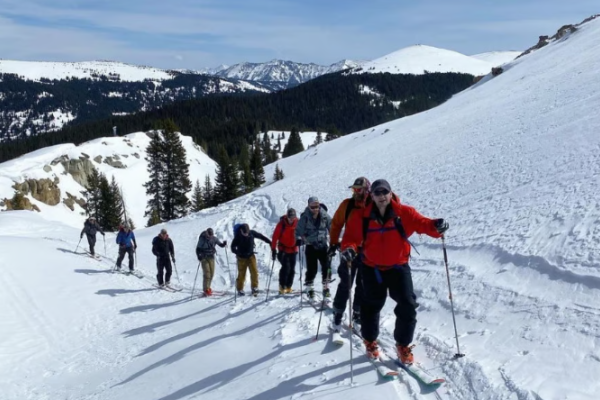 A group of people is skiing uphill on a snowy mountain slope surrounded by trees, under a clear blue sky.