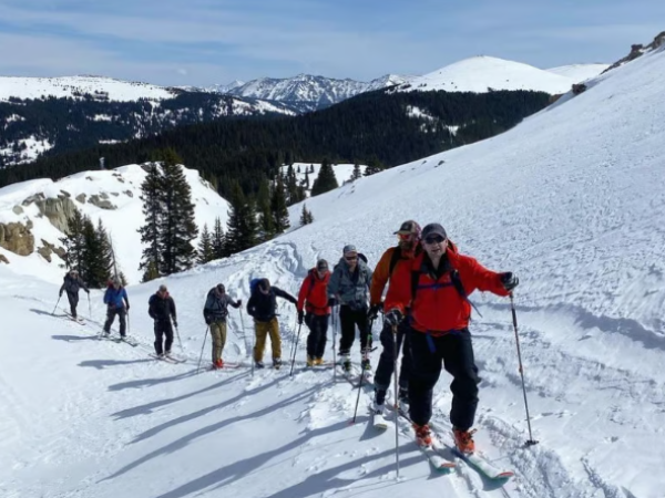 A group of people is skiing uphill on a snowy mountain slope surrounded by trees, under a clear blue sky.
