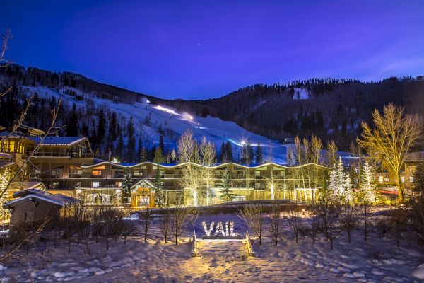 A snowy mountain resort at night with illuminated "VAIL" sign, surrounded by trees and buildings under a clear, deep blue sky.