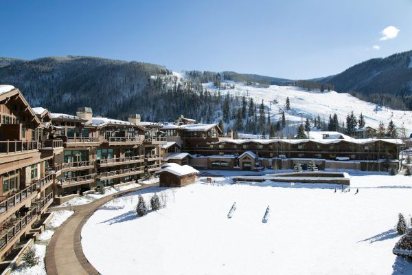 A snowy ski resort with wooden lodges and mountains in the background under a clear blue sky.