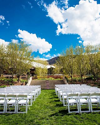 An outdoor venue with rows of white chairs set up on grass, surrounded by trees and under a blue sky with clouds.