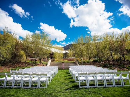 An outdoor venue with rows of white chairs set up on grass, surrounded by trees and under a blue sky with clouds.