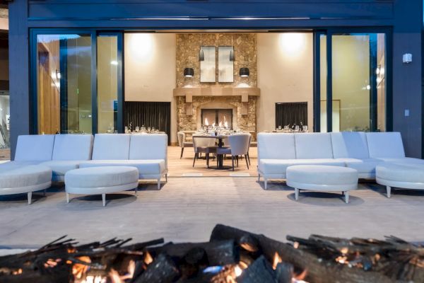 An outdoor seating area with modern furniture faces a large indoor fireplace, visible through a glass wall, with a fire pit in the foreground.