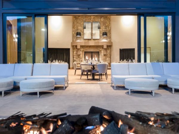 An outdoor seating area with modern furniture faces a large indoor fireplace, visible through a glass wall, with a fire pit in the foreground.