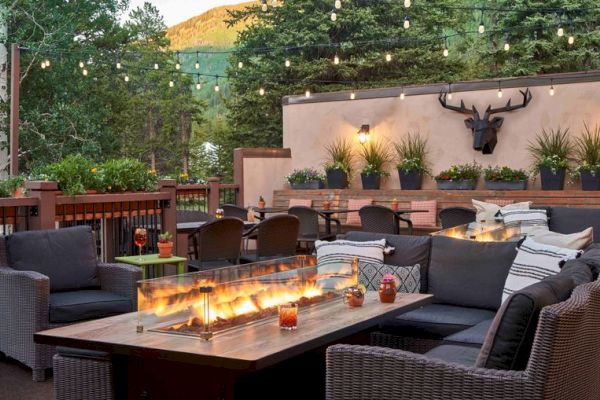 A cozy outdoor seating area with a long, rectangular fire pit table, surrounded by wicker furniture, string lights, and potted plants on a sunny day.