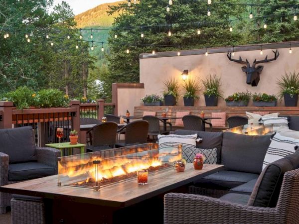 A cozy outdoor seating area with a long, rectangular fire pit table, surrounded by wicker furniture, string lights, and potted plants on a sunny day.