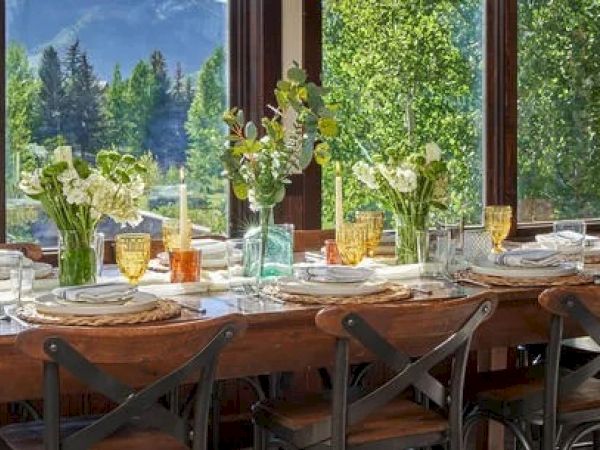 A dining table set for a meal, adorned with flowers and glassware, is situated in a room with large windows showing a scenic view of trees and mountains.