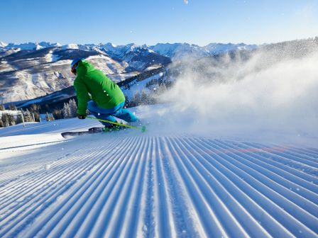 A skier in a green jacket and blue pants carves through fresh snow on a sunlit mountainous slope, creating a spray of powder.