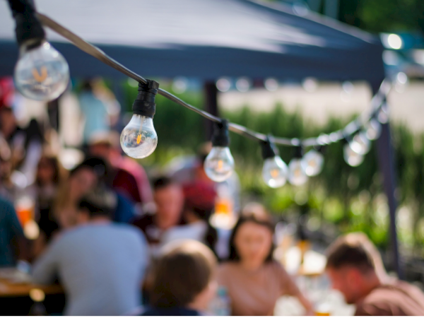 People sitting at an outdoor gathering under string lights; festive bulbs in focus while blurred guests chat and eat in the background.