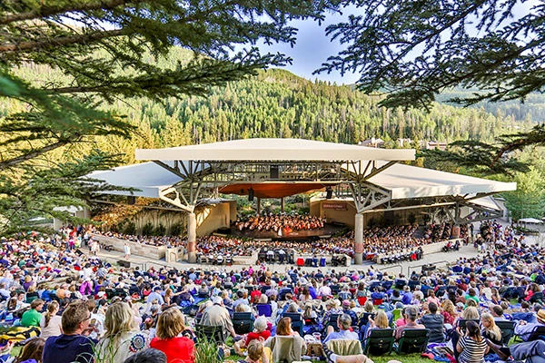 An outdoor concert with a seated audience under a canopy, surrounded by lush greenery and trees, set in a mountainous area.