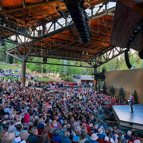 A large outdoor theater with a full audience under a wooden canopy, focused on a speaker on stage, in a scenic, wooded setting.