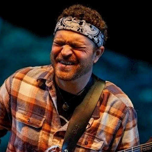 A person wearing a bandana and plaid shirt is playing a guitar with a focused expression, possibly during a live performance.