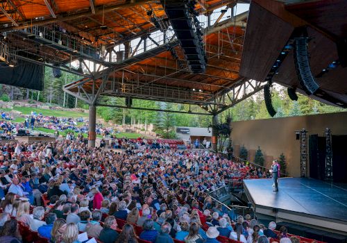 A large outdoor theater with a full audience under a wooden canopy, focused on a speaker on stage, in a scenic, wooded setting.