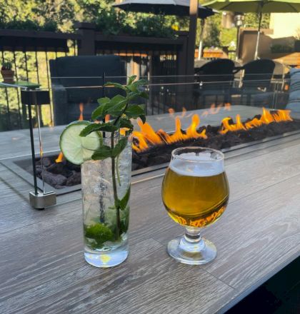 Two drinks, one with a lime and mint garnish, are on a table by a fire pit with outdoor seating in the background.