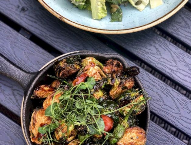 A skillet with roasted Brussels sprouts and greens, and a bowl of salad with lettuce and seeds are on a gray wooden surface.