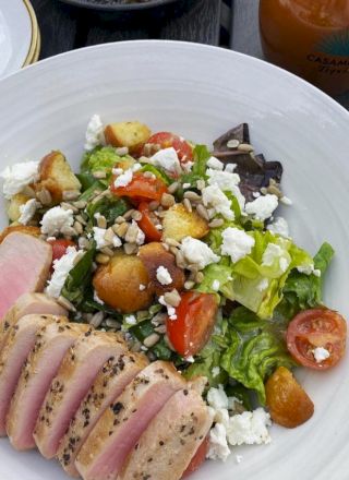 A salad with seared tuna, croutons, cherry tomatoes, feta, sunflower seeds, and greens is served in a white bowl.