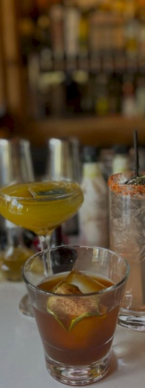 The image shows three cocktails on a bar counter, each with unique garnishes and styles, set against a blurred background of bottles.