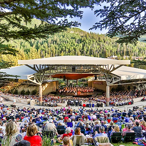 An outdoor concert with a seated audience under a canopy, surrounded by lush greenery and trees, set in a mountainous area.