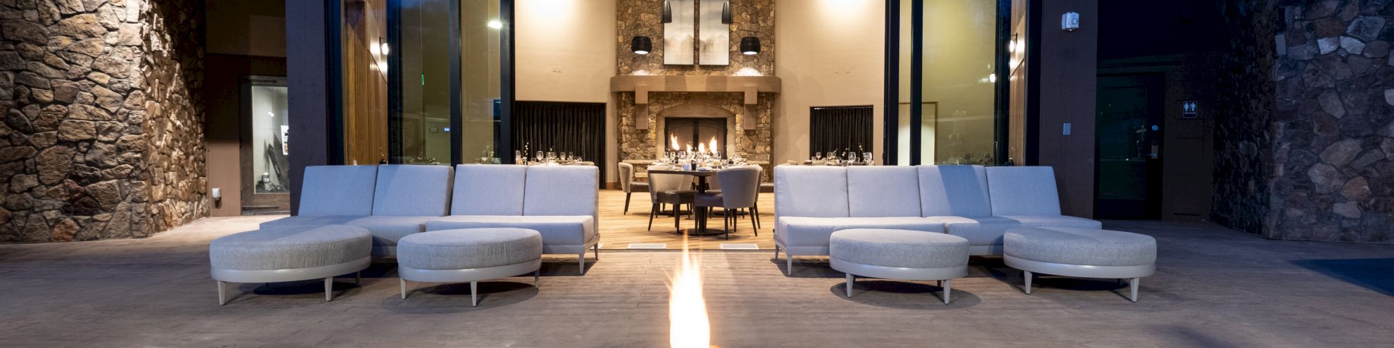 The image depicts a modern outdoor patio with a large fire pit in the foreground, surrounded by couches and a view of an interior room with a fireplace.