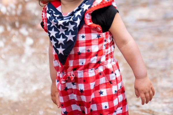 A child in a red-white checkered dress, heart-shaped sunglasses, and star-patterned scarf stands in shallow water.