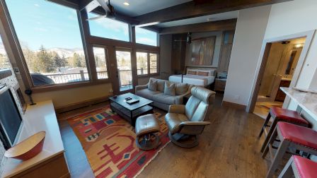 The image shows a cozy living area with a sofa, armchairs, TV, and colorful rug, alongside a bed and bar seating in a bright room.
