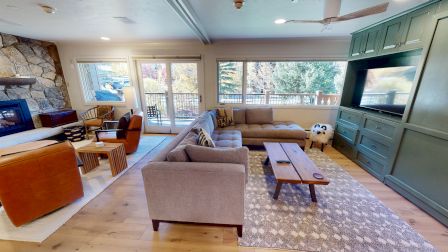 A cozy living room with a modern design, featuring sofas, a fireplace, large windows, and a TV. There's a patterned rug on the floor.