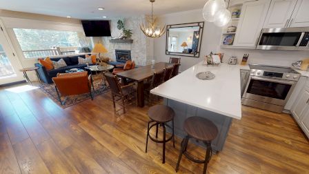 The image shows a modern open-plan living space with a kitchen, island seating, dining table, living area, and a wall-mounted TV.