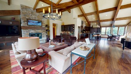 A spacious living room with a high ceiling, wooden beams, modern furniture, a stone fireplace, and open dining area in the background.