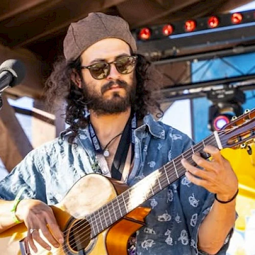A musician with curly hair and sunglasses plays an acoustic guitar on stage, wearing a patterned shirt and a hat, under bright lighting.