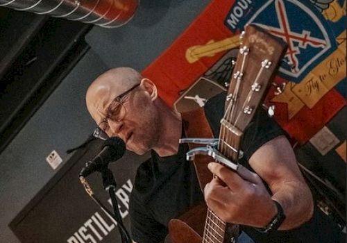 A person is playing an acoustic guitar and singing into a microphone indoors, with a colorful wall hanging in the background.