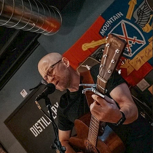 A person is playing an acoustic guitar and singing into a microphone indoors, with a colorful wall hanging in the background.